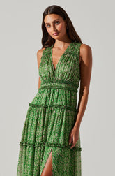 A woman is wearing a sleeveless, V-neck, green dress with a floral or abstract pattern. The dress features a gathered waist and tiered, ruffled sections, with a slit up one side revealing her leg. The fabric appears lightweight and semi-sheer.